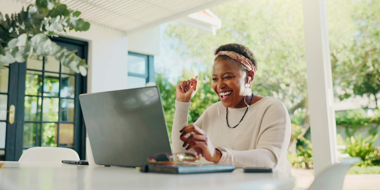 A smiling woman works from home outdoors, looking excitedly at her laptop screen while on a video call, representing someone working remotely for a flexible job.