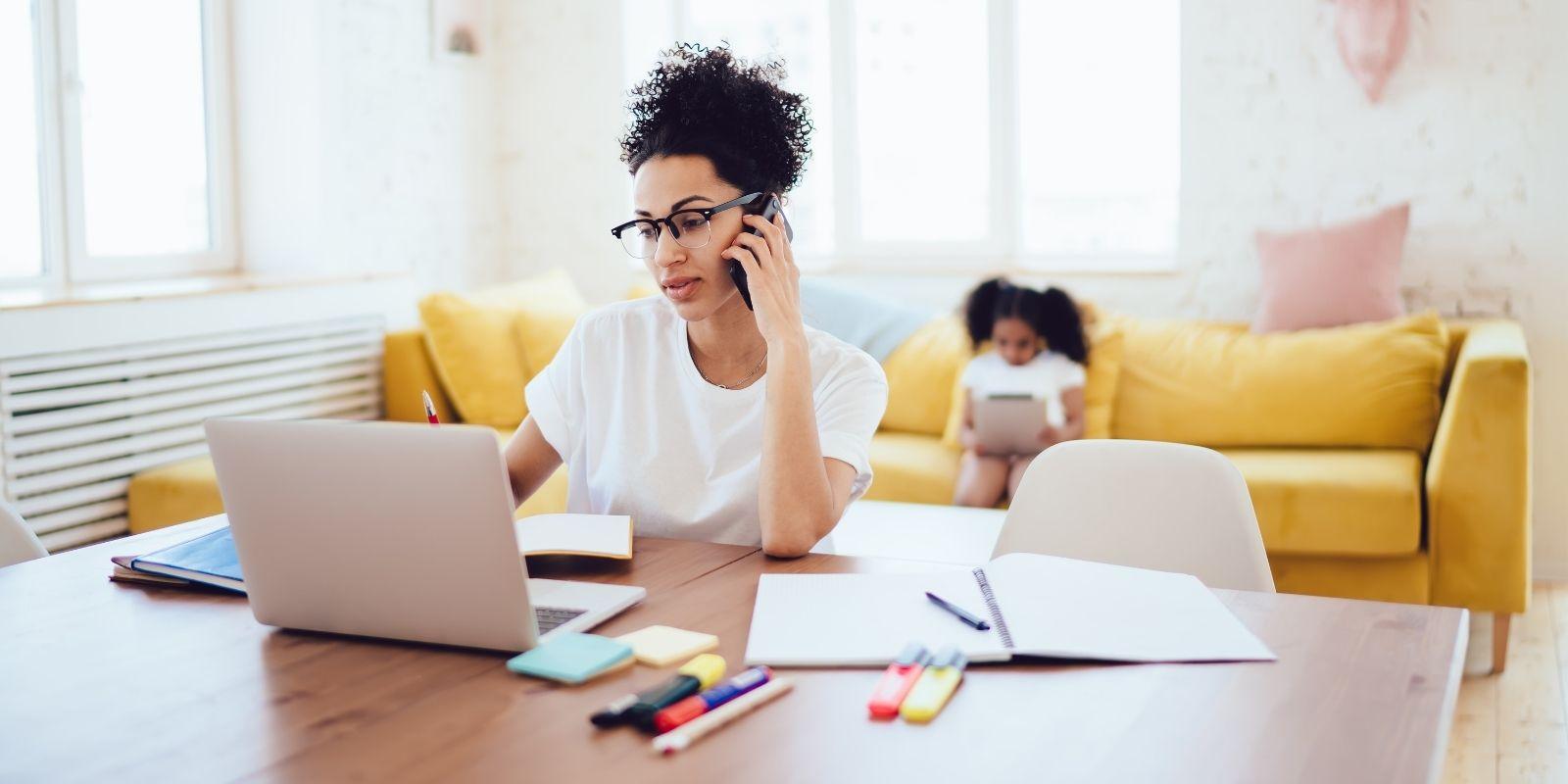 A woman works on a laptop and phone at home while a child sits on a couch in the background, suggesting a side hustle.