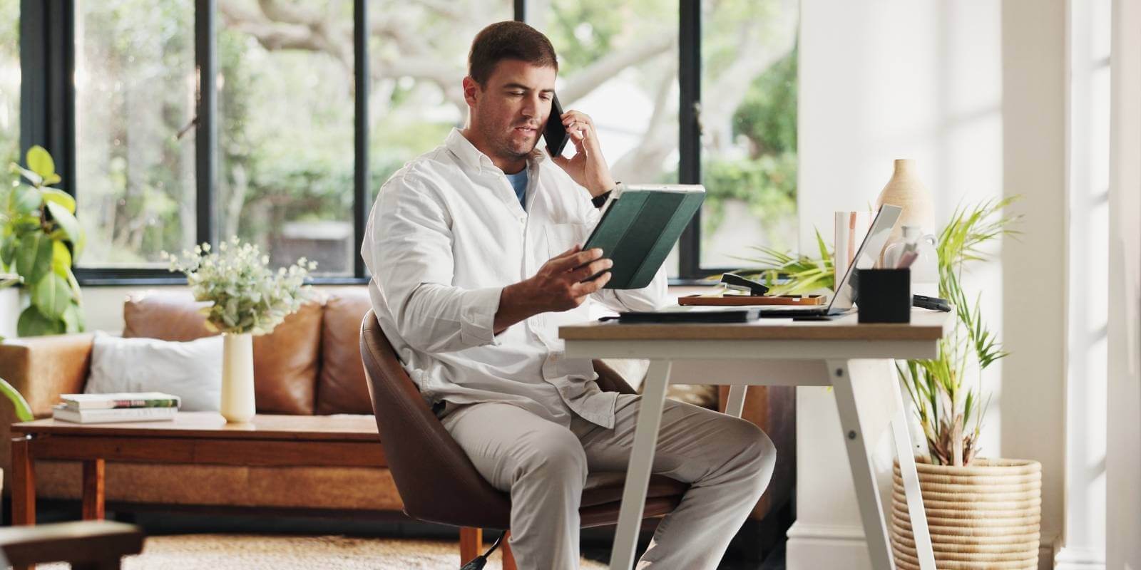 A man sits at a desk in a home office, talking on a smartphone while looking at a tablet, representing someone working a remote admin job.