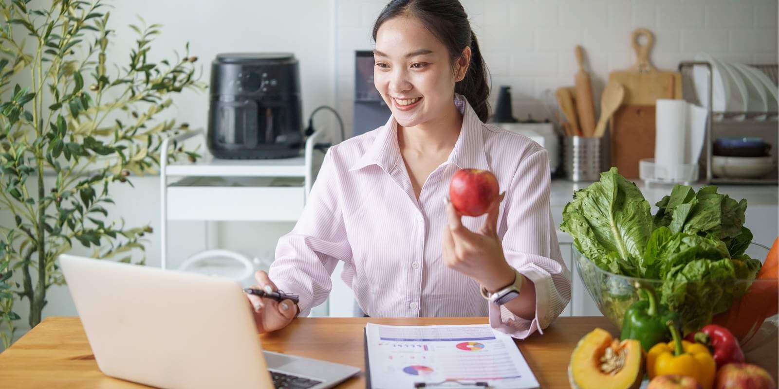 A smiling woman sits at a table with a laptop on a video call, holding an apple while reviewing charts near a bowl of fresh vegetables, representing a remote health coach.